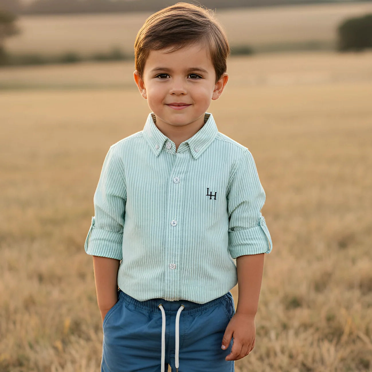 A young boy wearing a light blue-green and white vertically striped button-down shirt with a small black embroidered logo on the chest, paired with blue drawstring pants, standing in a dry grass field.