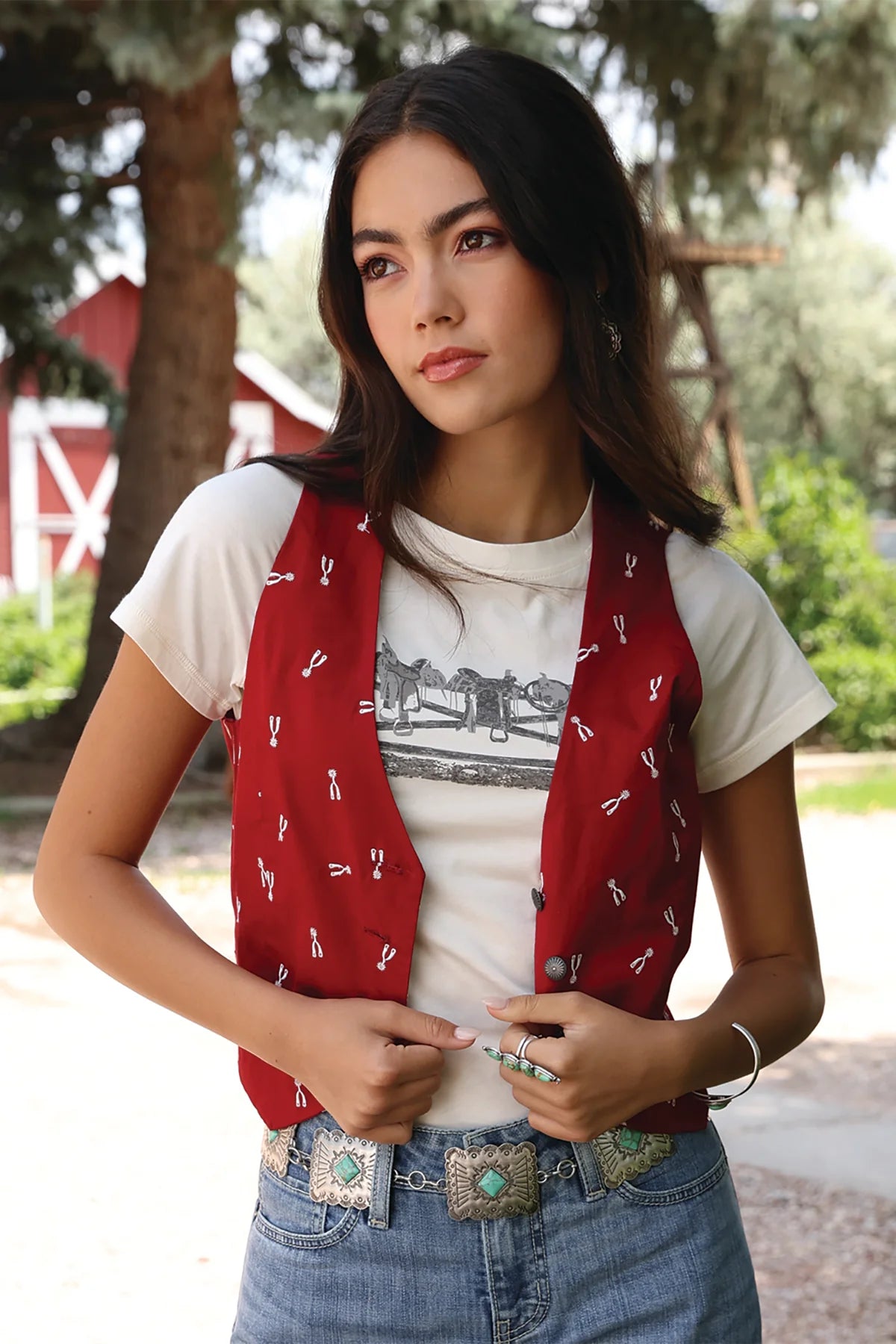 A woman wearing a red western-style vest with white embroidered details over a white graphic t-shirt and blue jeans with a western belt, standing outdoors in front of trees and a red barn.