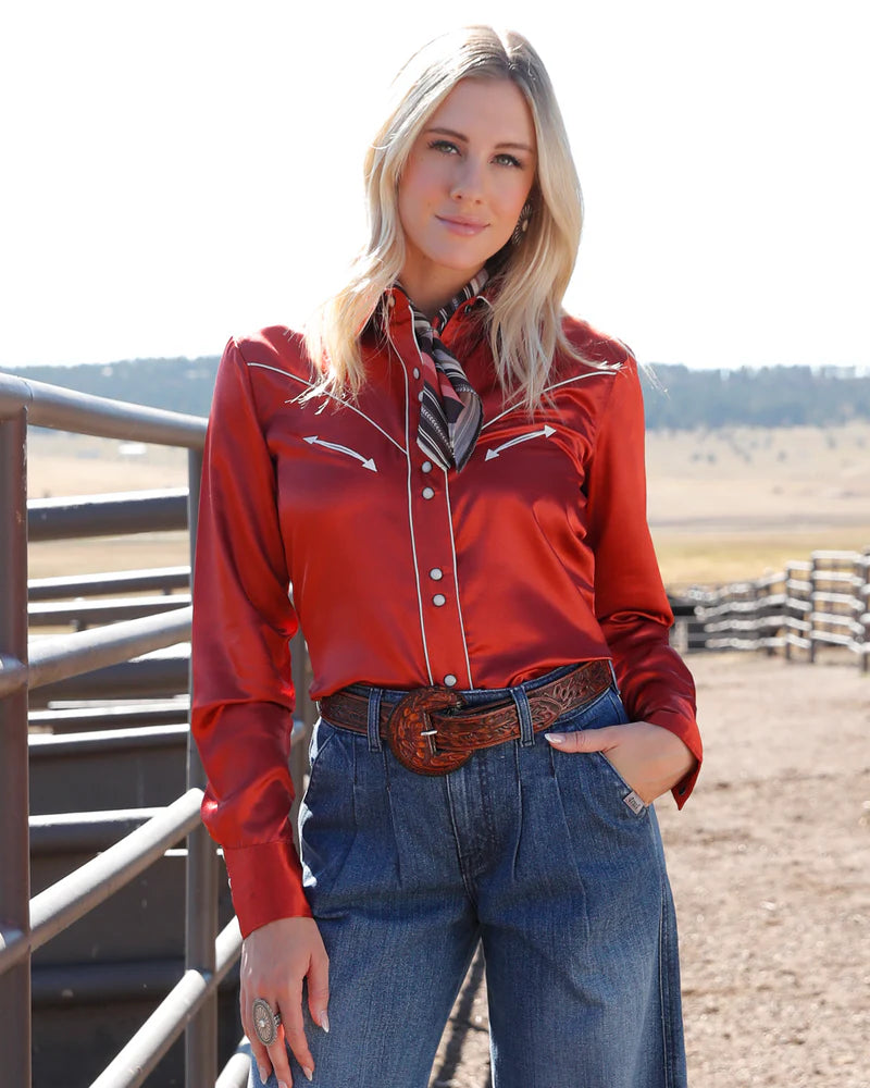 Woman wearing a red blouse and blue jeans with a scarf, standing in an outdoor setting.