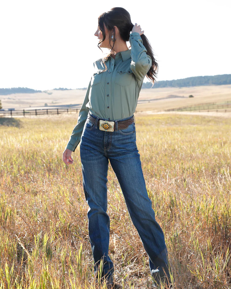 Woman in a green shirt and blue jeans standing in a field with a belt buckle.
