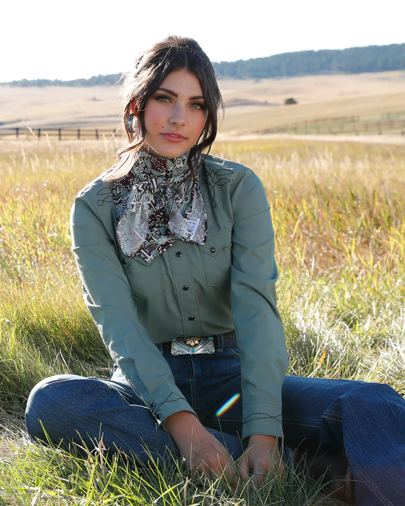 Woman sitting in a field wearing a green shirt and blue jeans with a patterned scarf.