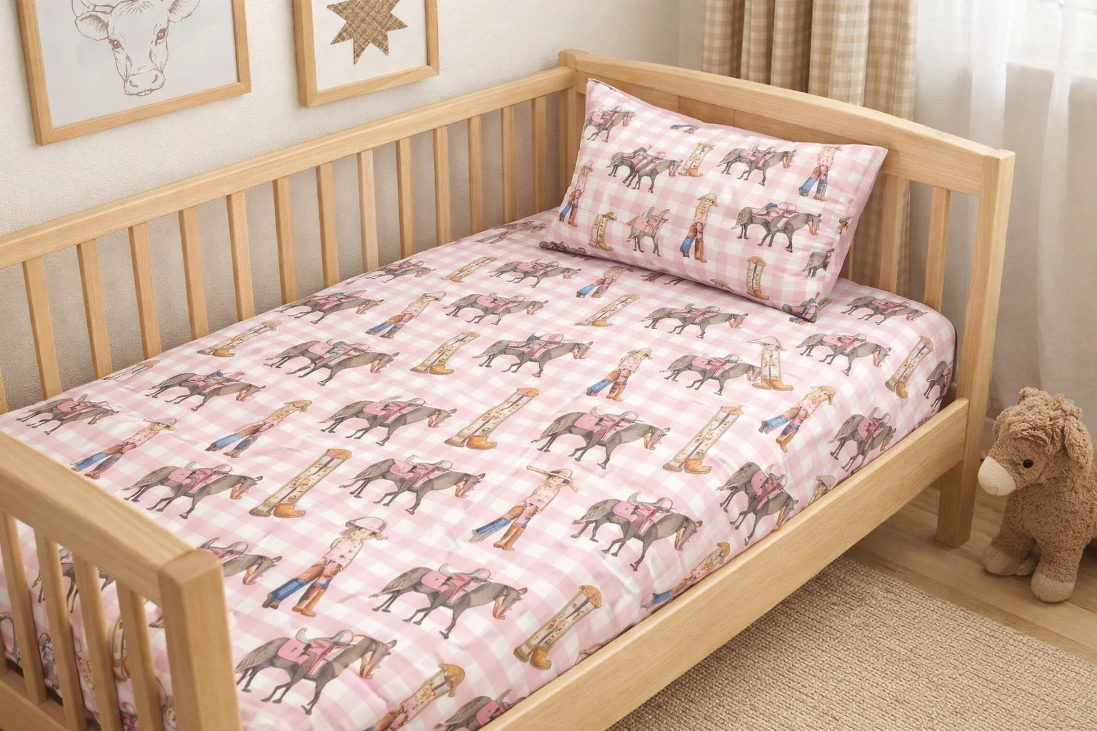 Wooden crib with pink bedding featuring animal patterns, a teddy bear, and framed pictures in the background.