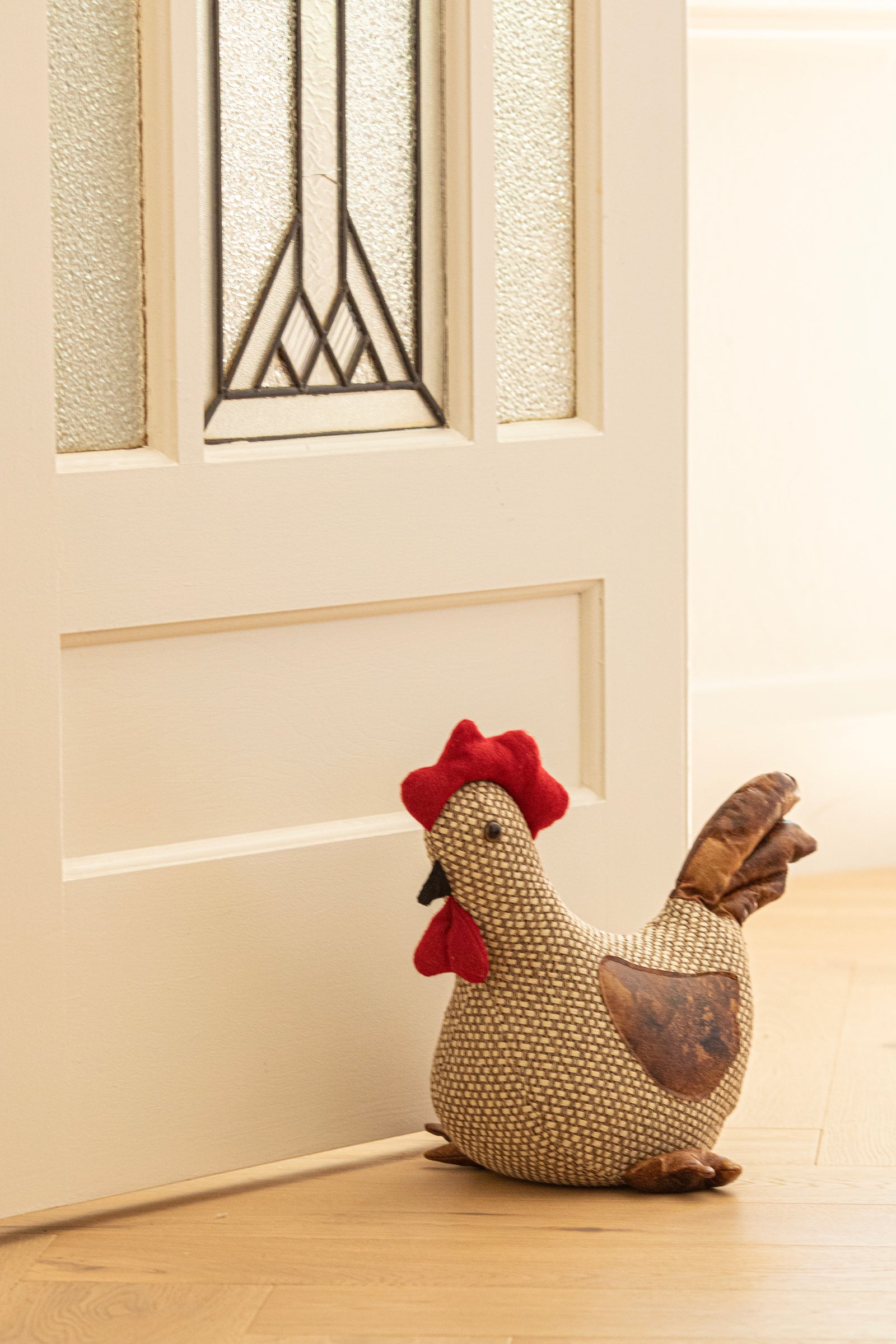 A woven chicken-shaped door stopper with a red comb and wattle, resting on a light wood floor next to a cream-colored paneled door.