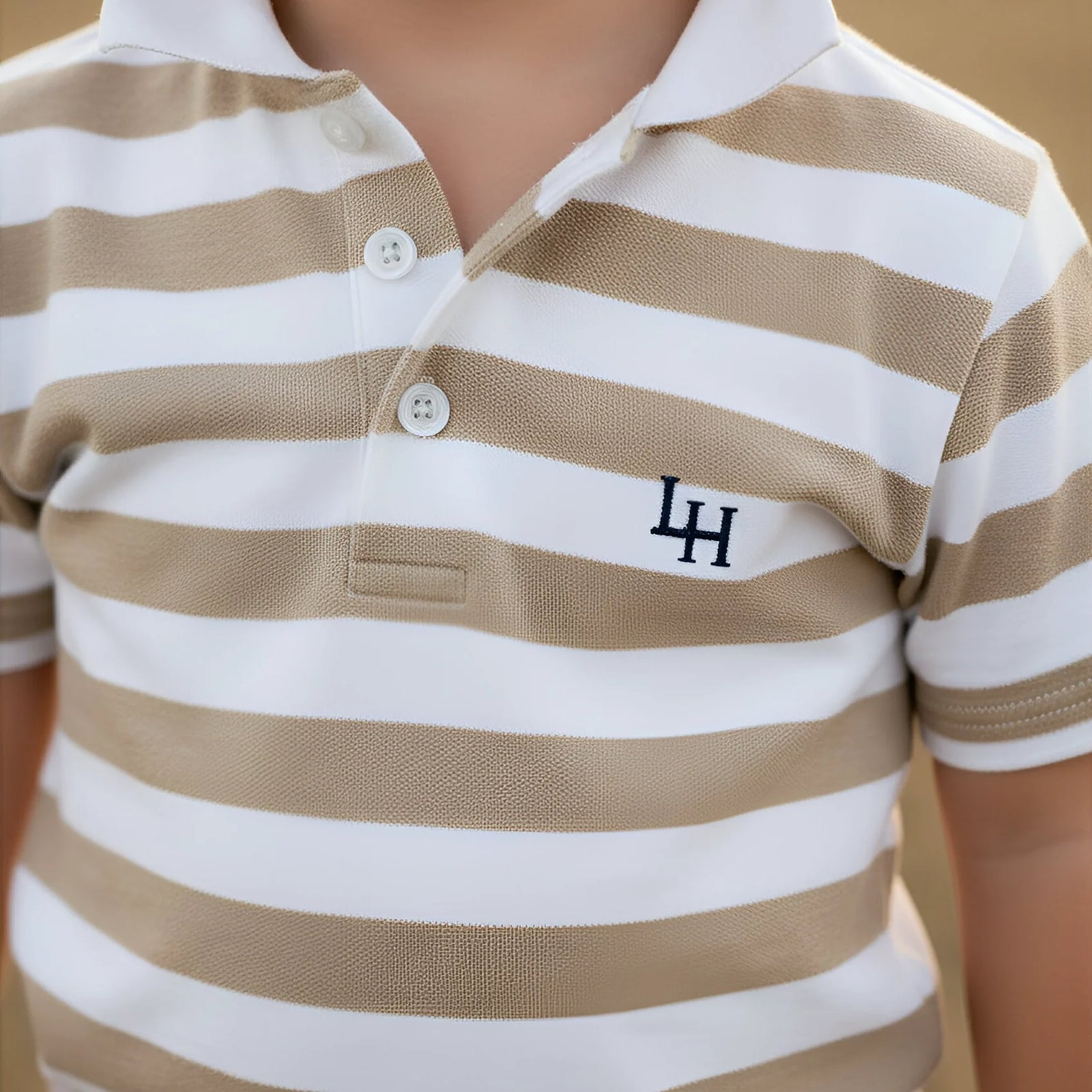 A close-up of a child wearing a short-sleeve polo shirt with beige and white horizontal stripes. The shirt features a white embroidered 'LA' logo on the left chest.