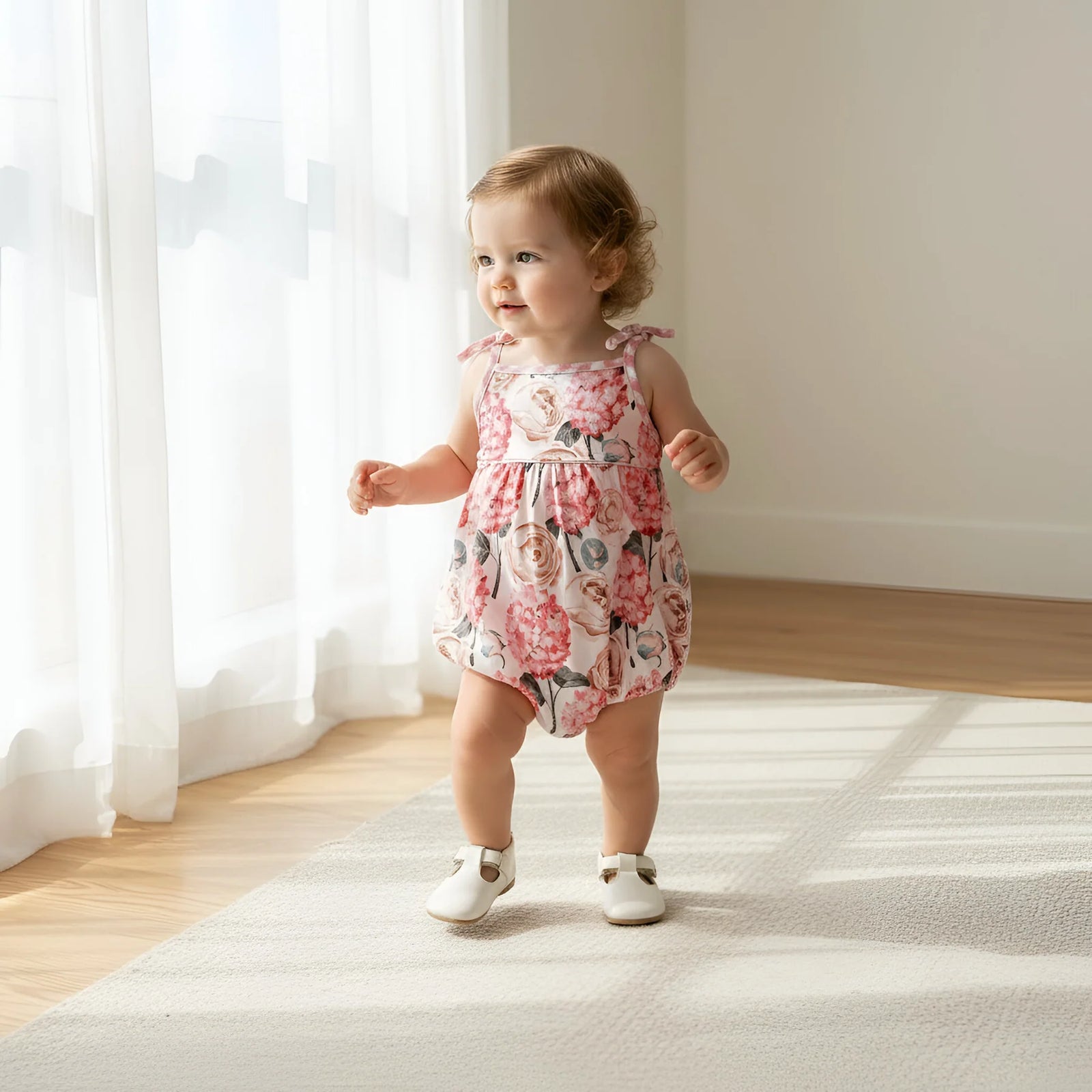 A toddler girl wearing a sleeveless floral romper and white shoes stands on a light rug in a bright room with sheer curtains and wooden floors.