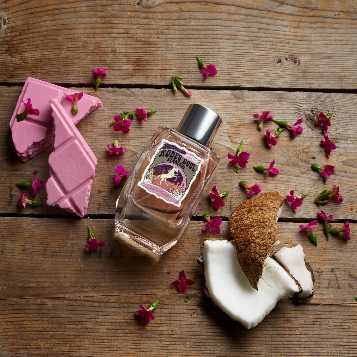 Perfume bottle with pink chocolate and coconut on a wooden surface with flowers.