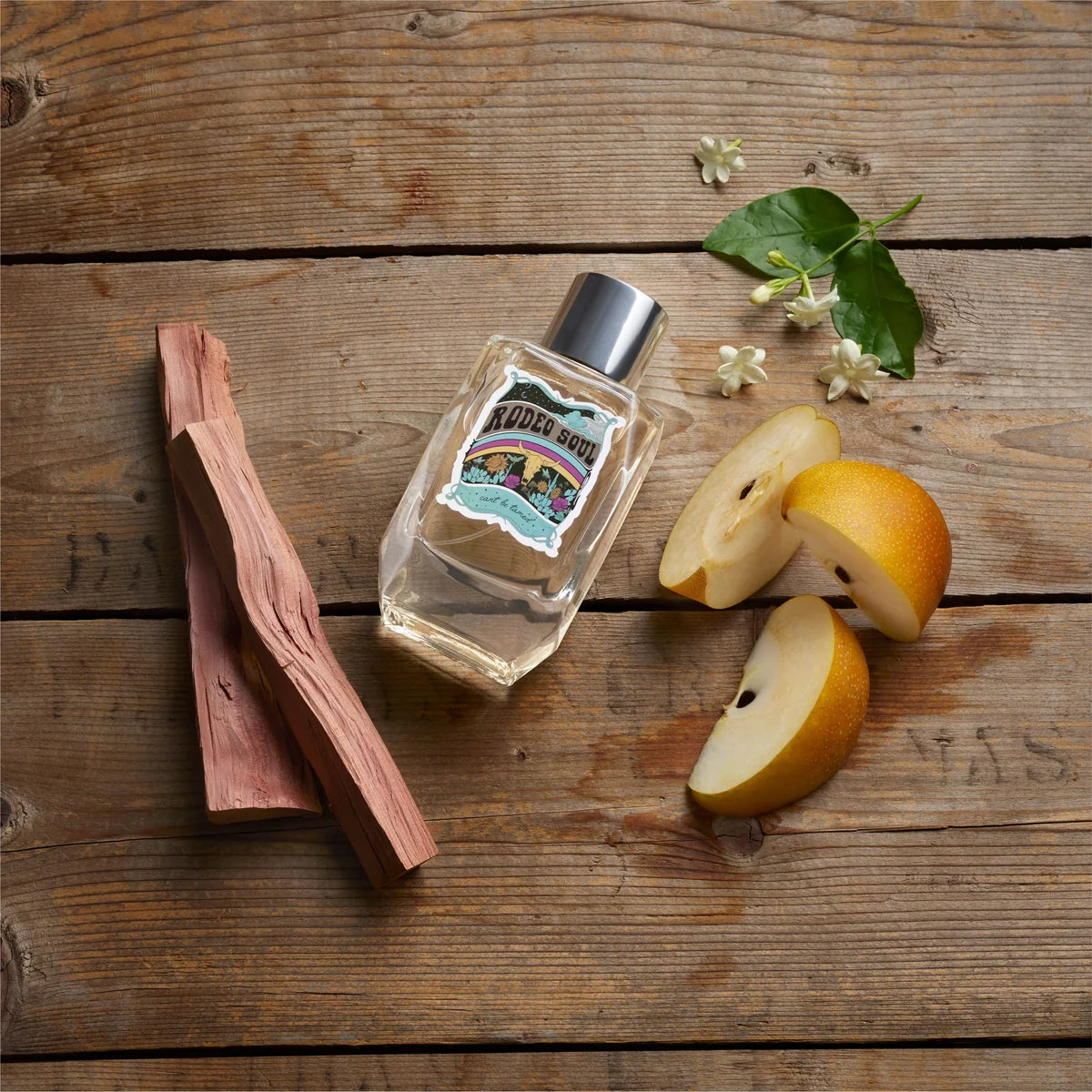 A clear glass bottle of scented soap with a silver cap and a decorative front label, styled on a rustic wooden surface with sliced pears, wood chips, and small white flowers.