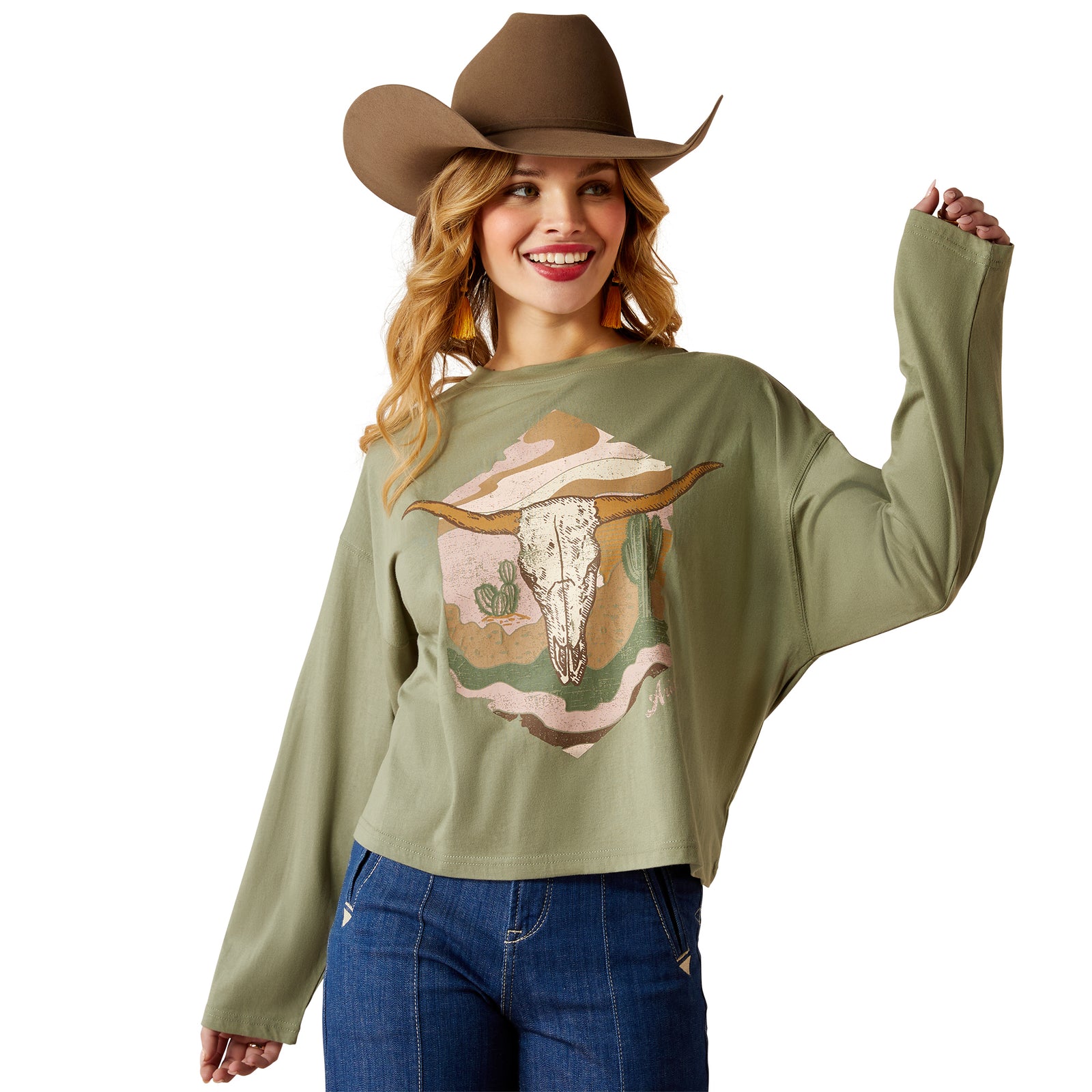 Ariat Women's Wanderer L/S T-Shirt. A woman wearing a long-sleeved green t-shirt with a graphic print of a cow's skull, paired with a brown cowboy hat and blue jeans.