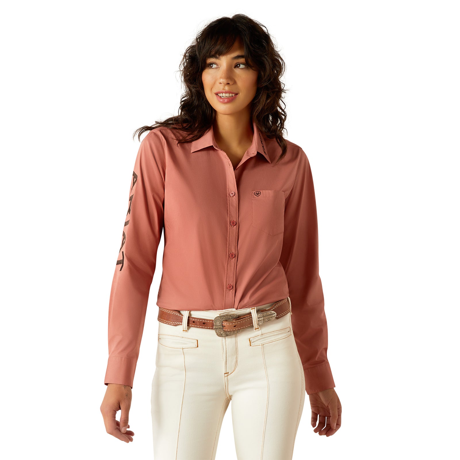 Ariat Women's Wrinkle Resistant Team Kirby L/S Western Shirt. A woman wearing a long-sleeved pink western shirt with subtle pattern detailing on the sleeves, paired with light-colored pants.
