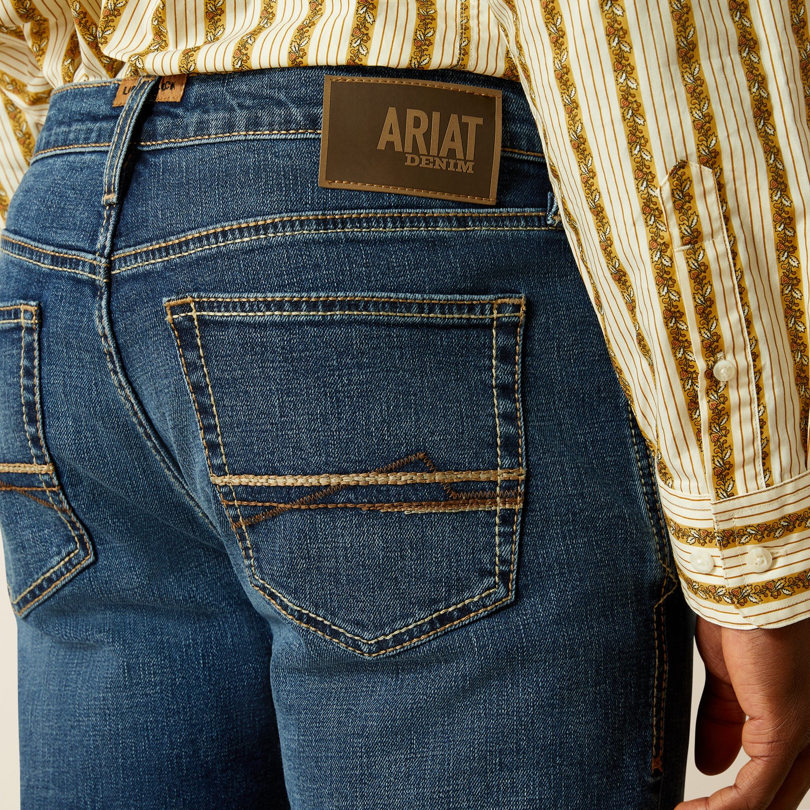 Ariat Men's Samuel M8 Straight Leg Jean. Close-up of blue jeans with Ariat denim label and striped shirt.