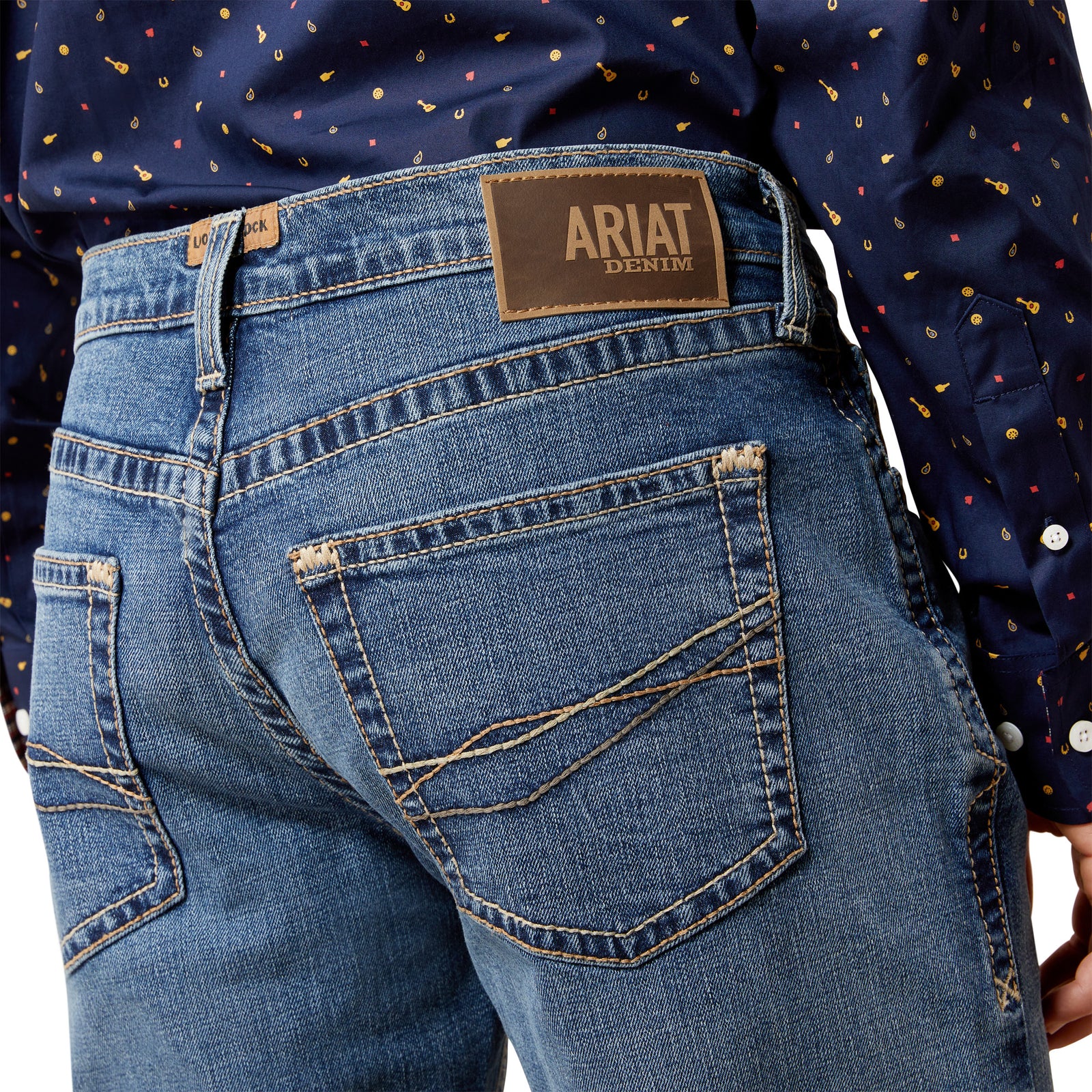 Close-up of blue jeans with Ariat denim label, worn by a person in a patterned shirt.