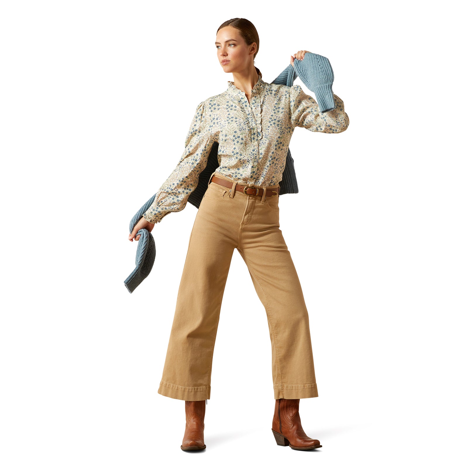 Woman wearing a patterned blouse, beige pants, and brown boots holding a blue jacket against a white background