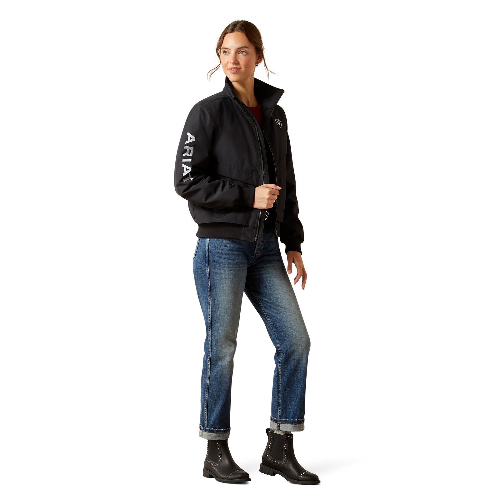 Woman wearing a black jacket with 'ARIAT' branding, blue jeans, and black boots on a white background
