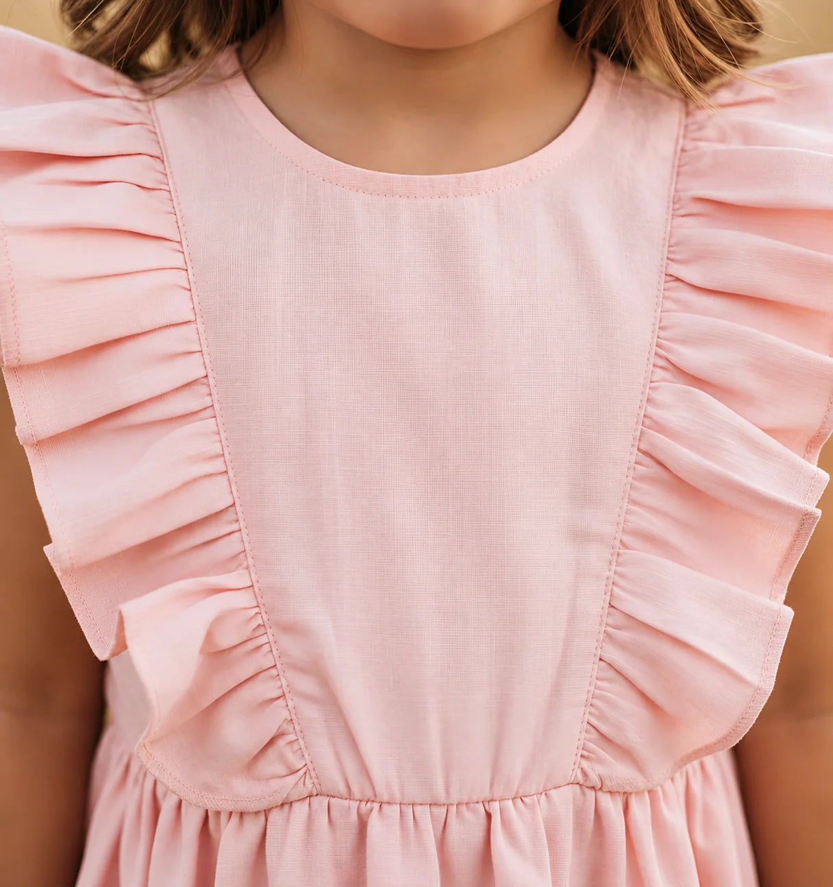 Close-up of a child wearing a light pink sleeveless dress with ruffled shoulder details and a gathered waist seam.