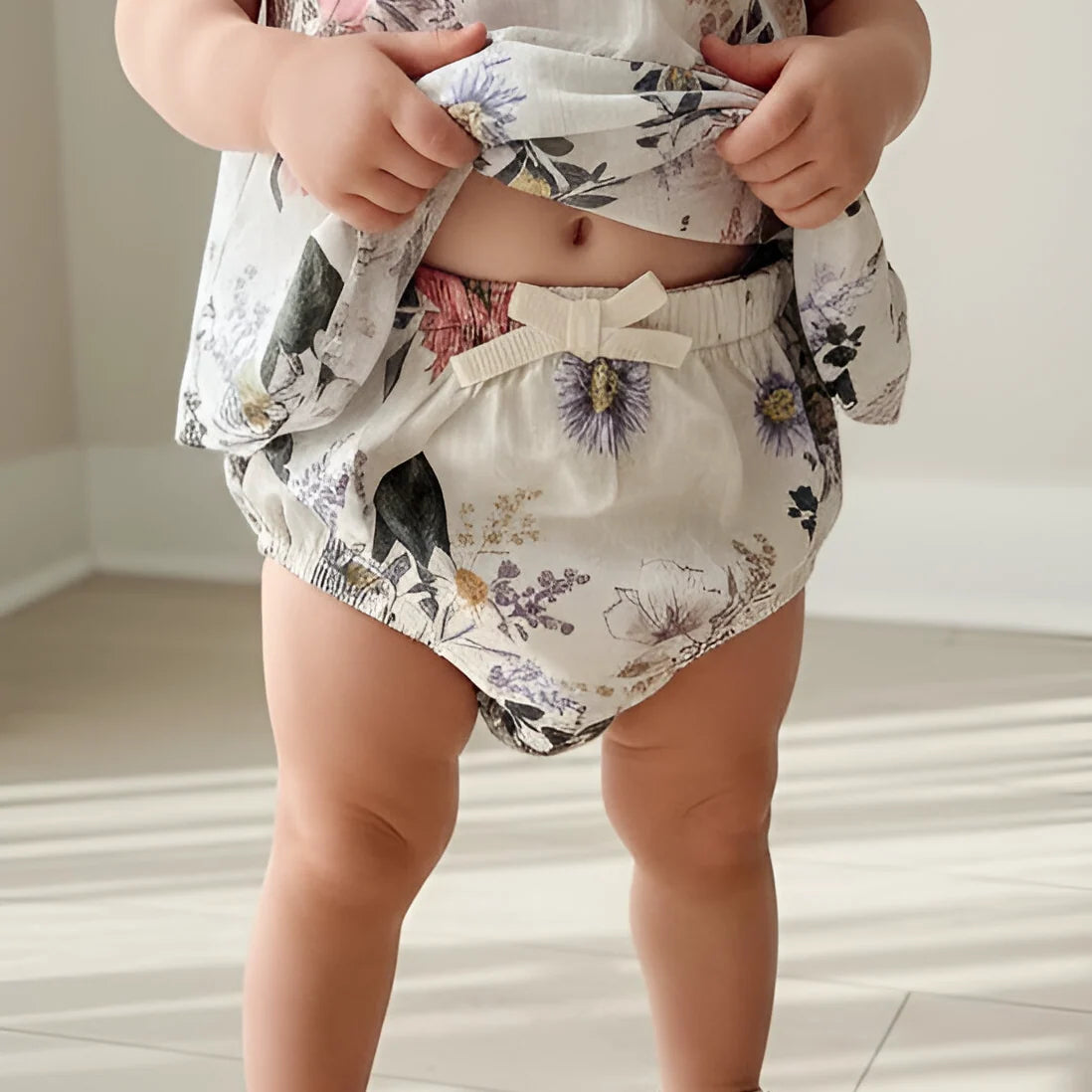 A close-up of a toddler wearing floral bloomers with a white elastic waistband and bow, holding up the matching top to show the fabric print.