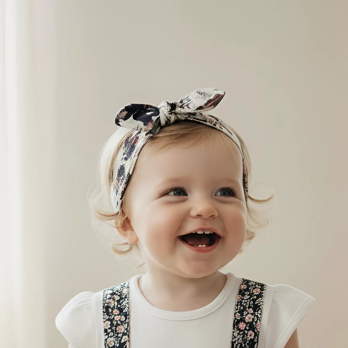 Baby wearing a white onesie with floral suspenders and a headband, standing against a light curtain background.