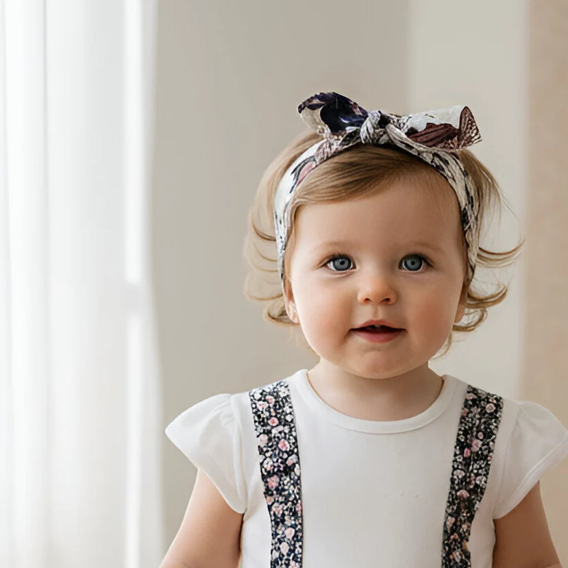 A toddler girl with light brown hair wearing a white t-shirt and floral patterned suspenders, accessorized with a matching floral bow headband, photographed in a bright, soft-focused indoor setting.