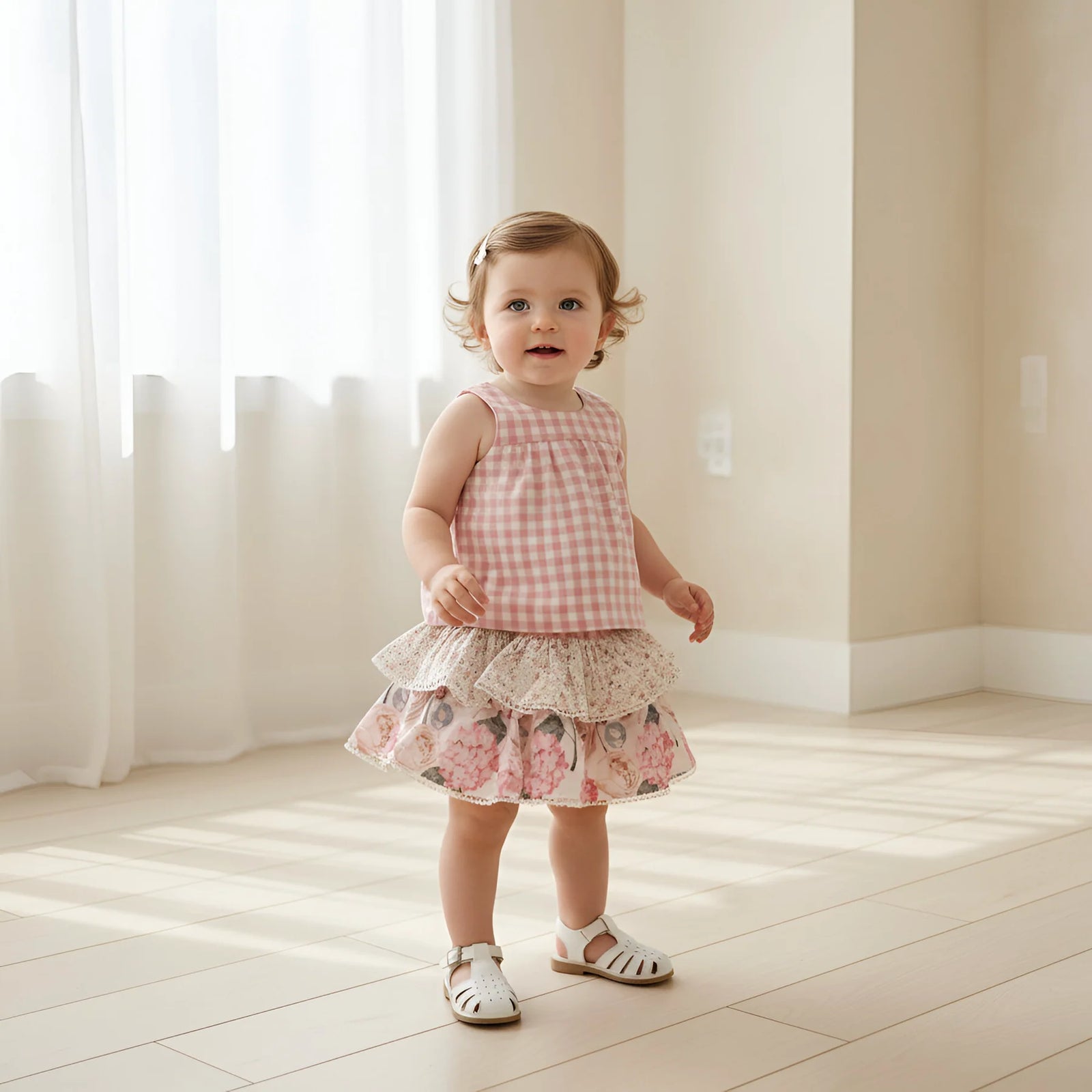 A toddler girl stands in a bright room with sunlight and sheer curtains, wearing a sleeveless pink-and-white gingham top and a tiered floral skirt, paired with white sandals.