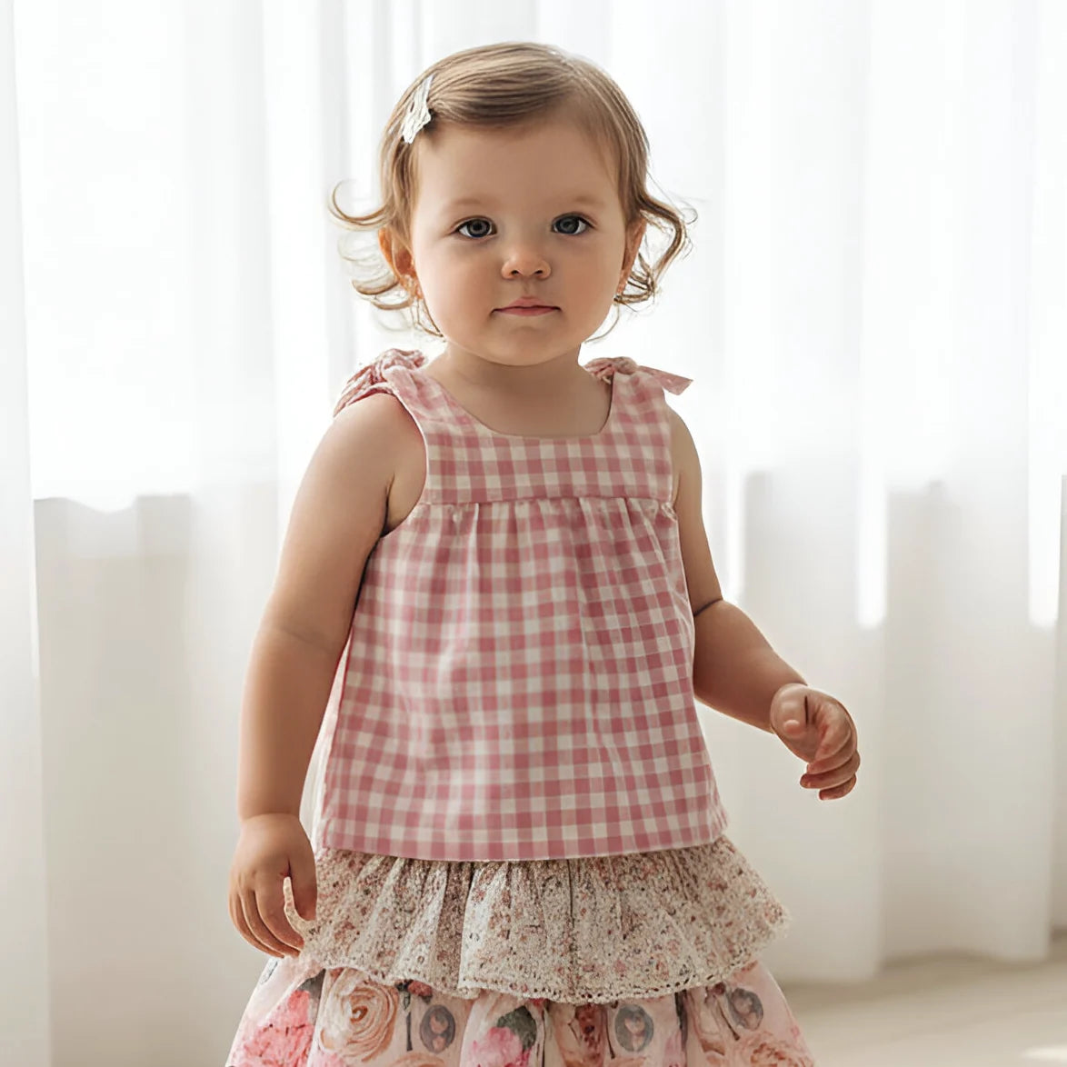 Young girl wearing a pink checkered top and floral skirt against a white curtain background