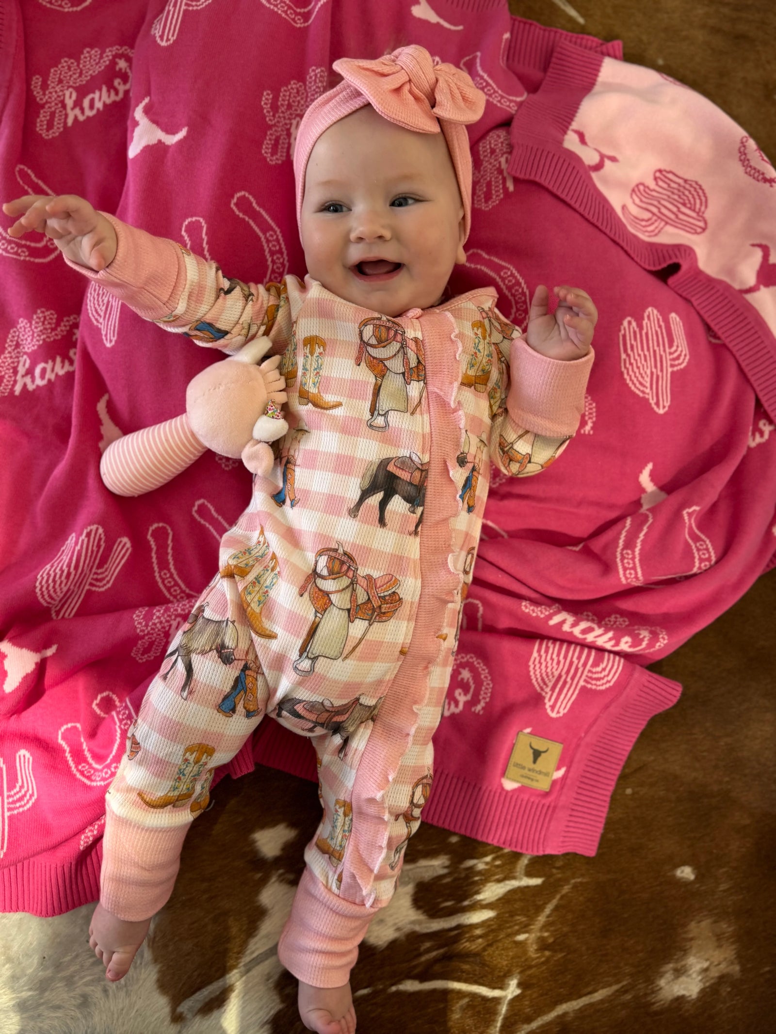 A baby wearing a pink and white gingham footless one-piece pajama with cowboy and cow print, matching pink booties, and a pink bow headband, lying on a pink blanket with a cowhide rug underneath.