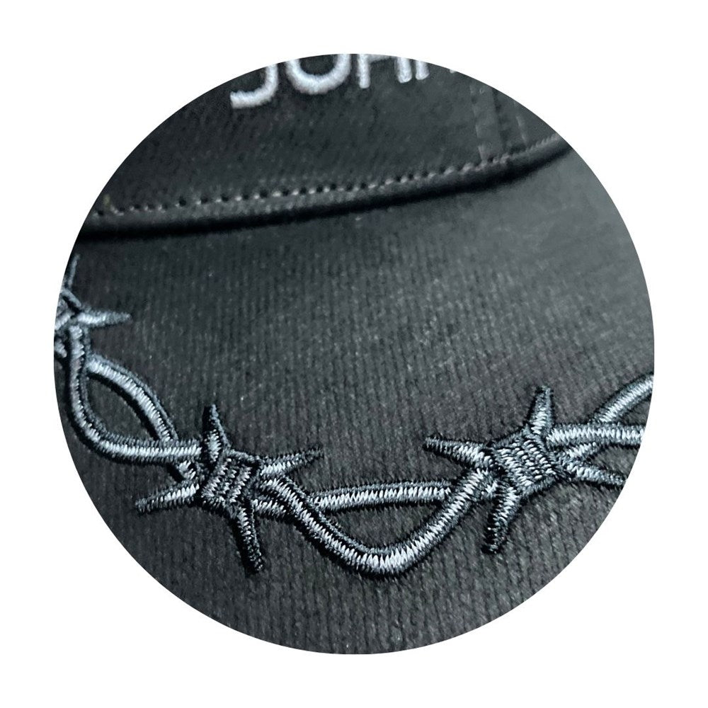 Close-up of a black baseball cap featuring detailed silver-gray embroidered barbed wire across the front panel, with a white embroidered letter 'J' partially visible at the top.