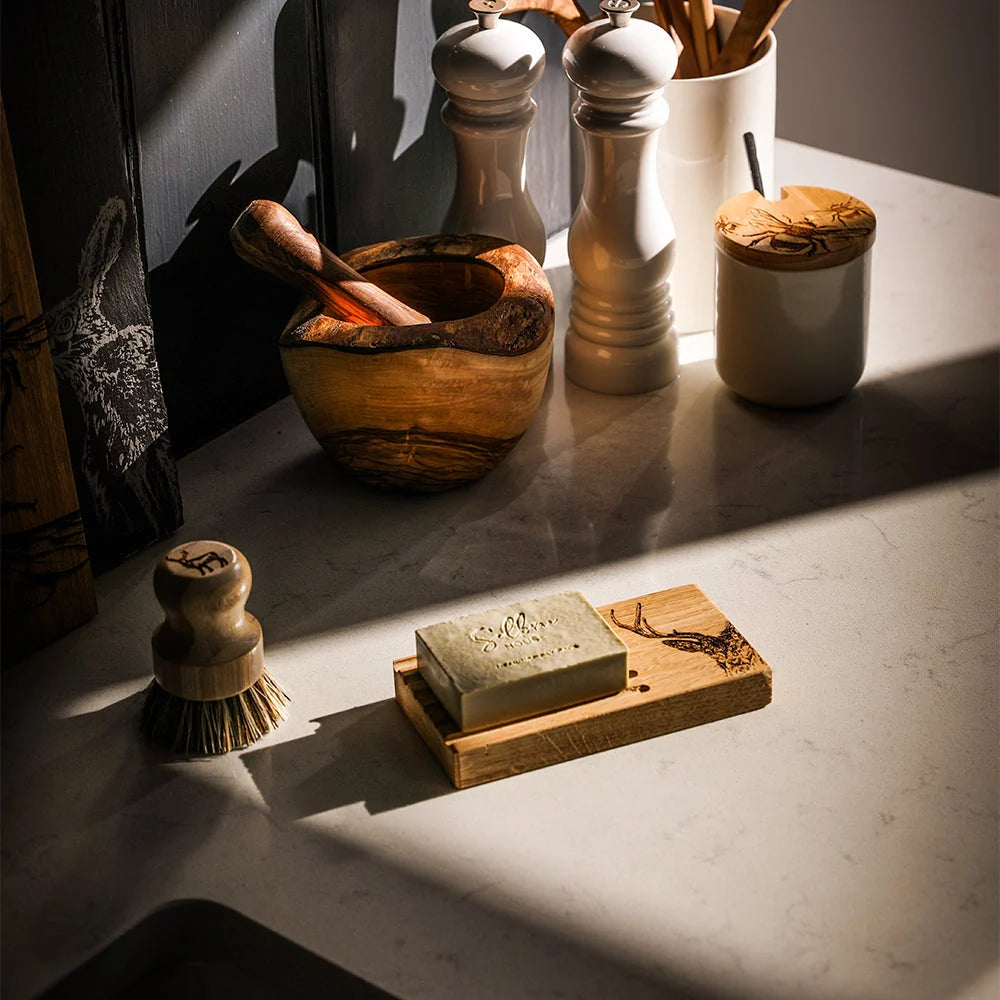 A bar of soap in a wooden soap dish sits on a light stone countertop in warm, directional sunlight, surrounded by kitchen items including a wooden bowl with a pestle, white salt and pepper mills, and a small brush.