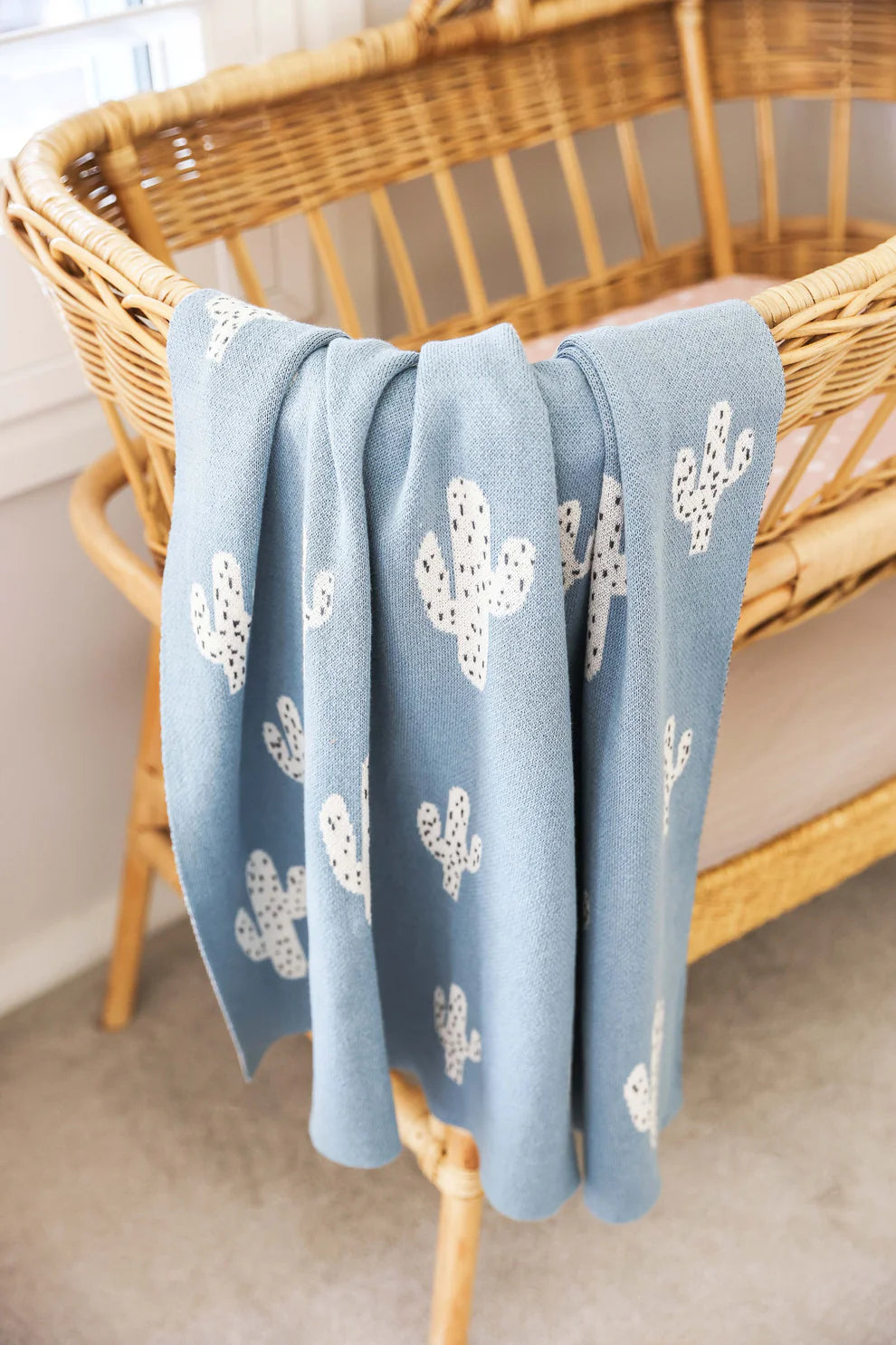 Alimrose Cactus Baby Blanket Blue. Blue blanket with white cactus patterns draped over a wicker chair.