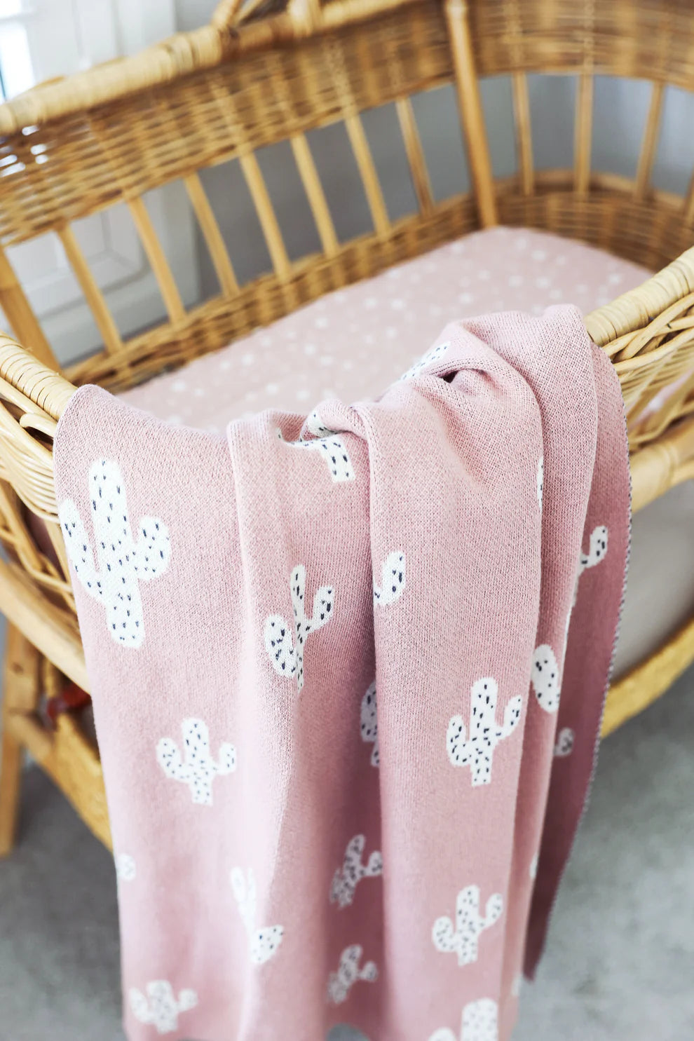 Alimrose Cactus Baby Blanket Petal. Pink blanket with white floral patterns draped over a wicker chair.