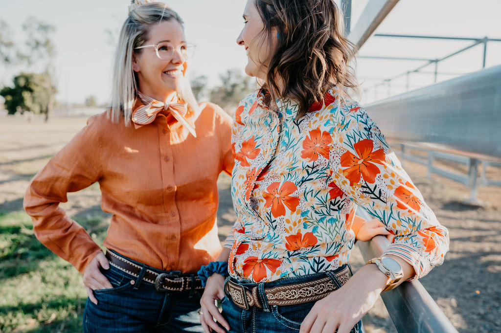 Two women standing outdoors, one wearing an orange leather jacket and the other in a floral blouse.