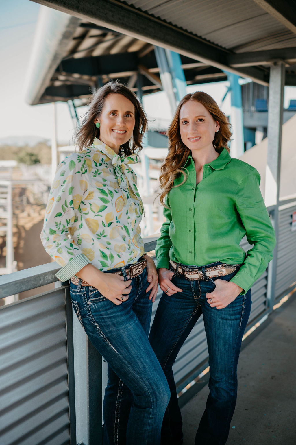 Two women standing side by side on a metal platform, wearing patterned shirts and jeans.