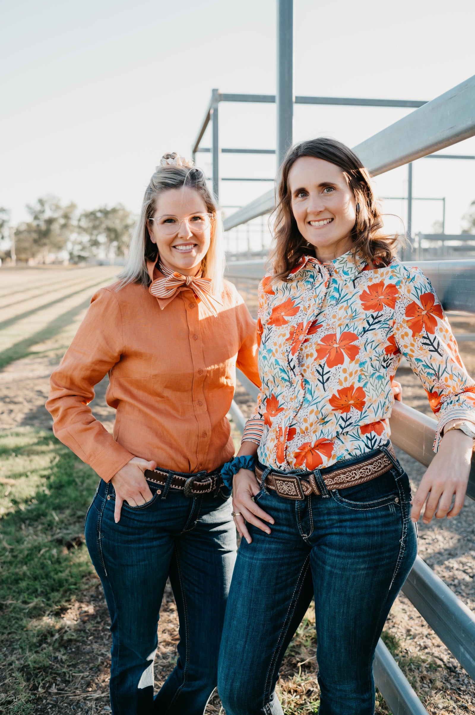 Two women standing outdoors near a metal fence, wearing colorful shirts and jeans.