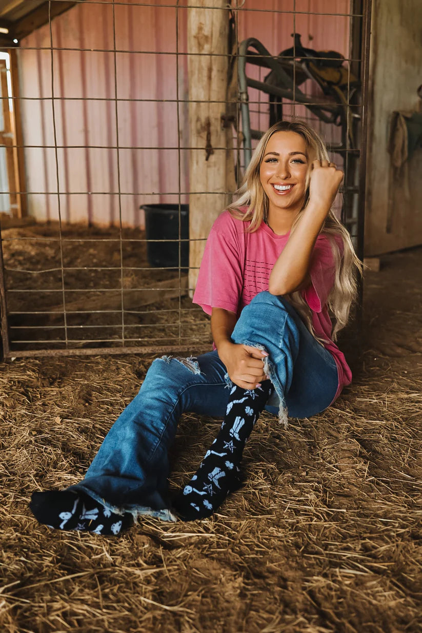 Lucky Chuck Get Western Performance Socks. Woman sitting on hay in a barn wearing a pink shirt and blue jeans.