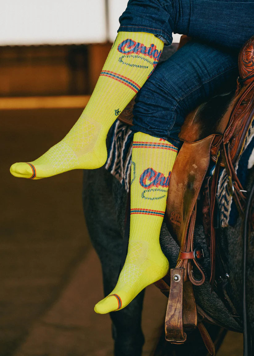 Lucky Chuck Cactus Alley Chicken Roping Performance Socks. Person wearing bright yellow socks with a brand logo, sitting on a horse.