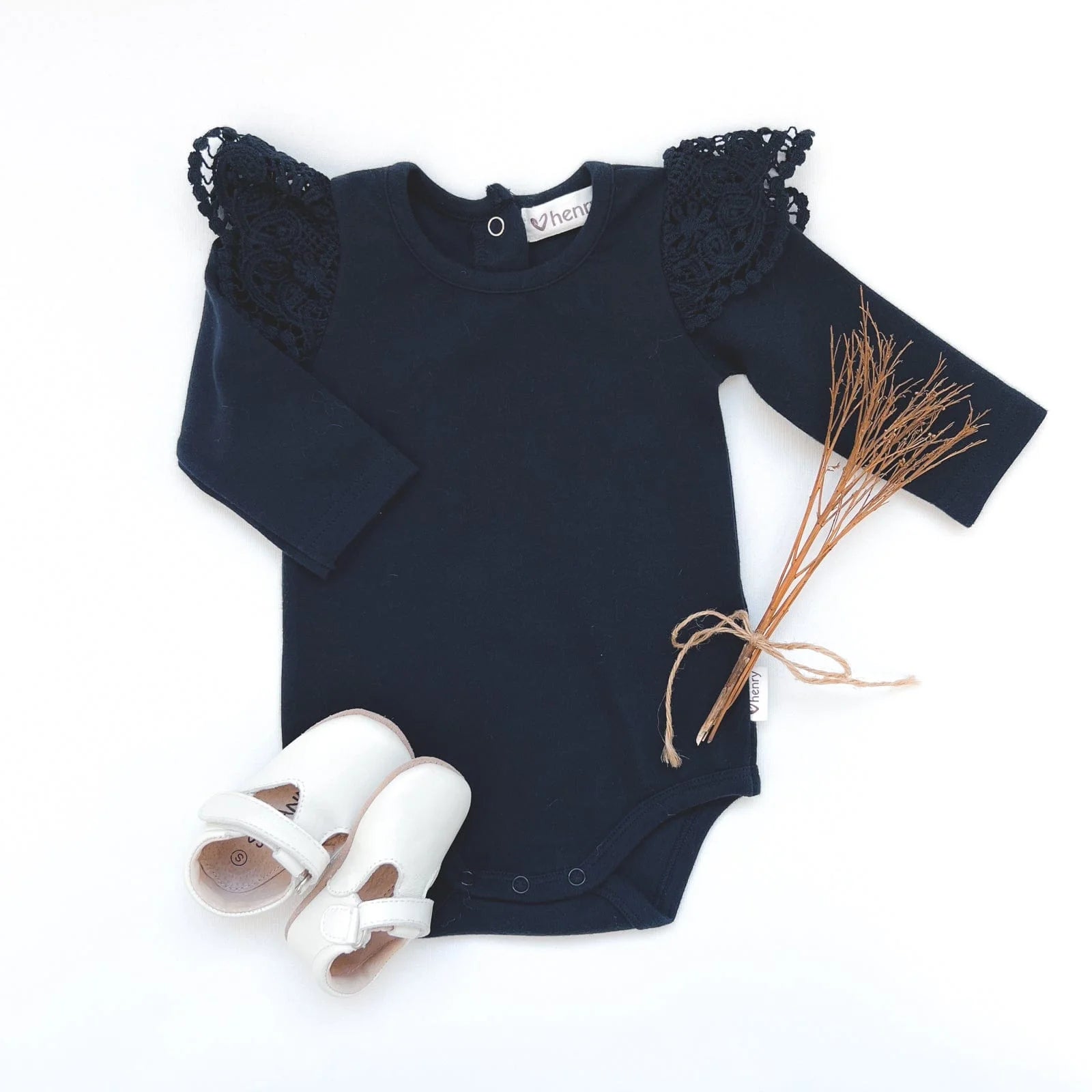 Love Henry Baby Girl's Lace Sleeve Onesie. Navy baby onesie with lace sleeves and a bias bound crew neck, accompanied by a pair of white shoes and a decorative element.