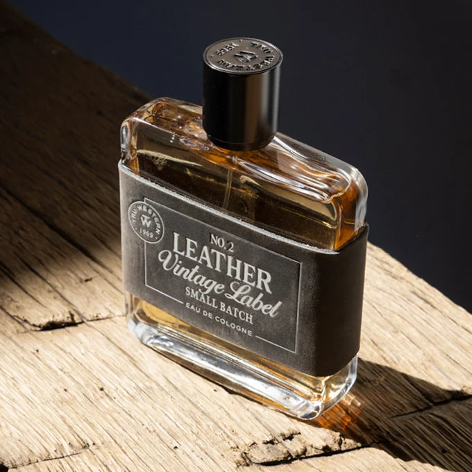 A bottle of No. 2 Leather Vintage Label Small Batch Eau de Cologne, featuring a clear glass bottle filled with amber liquid, a black cap, and a black paper label wrapped around the middle, resting on a rustic wooden surface.