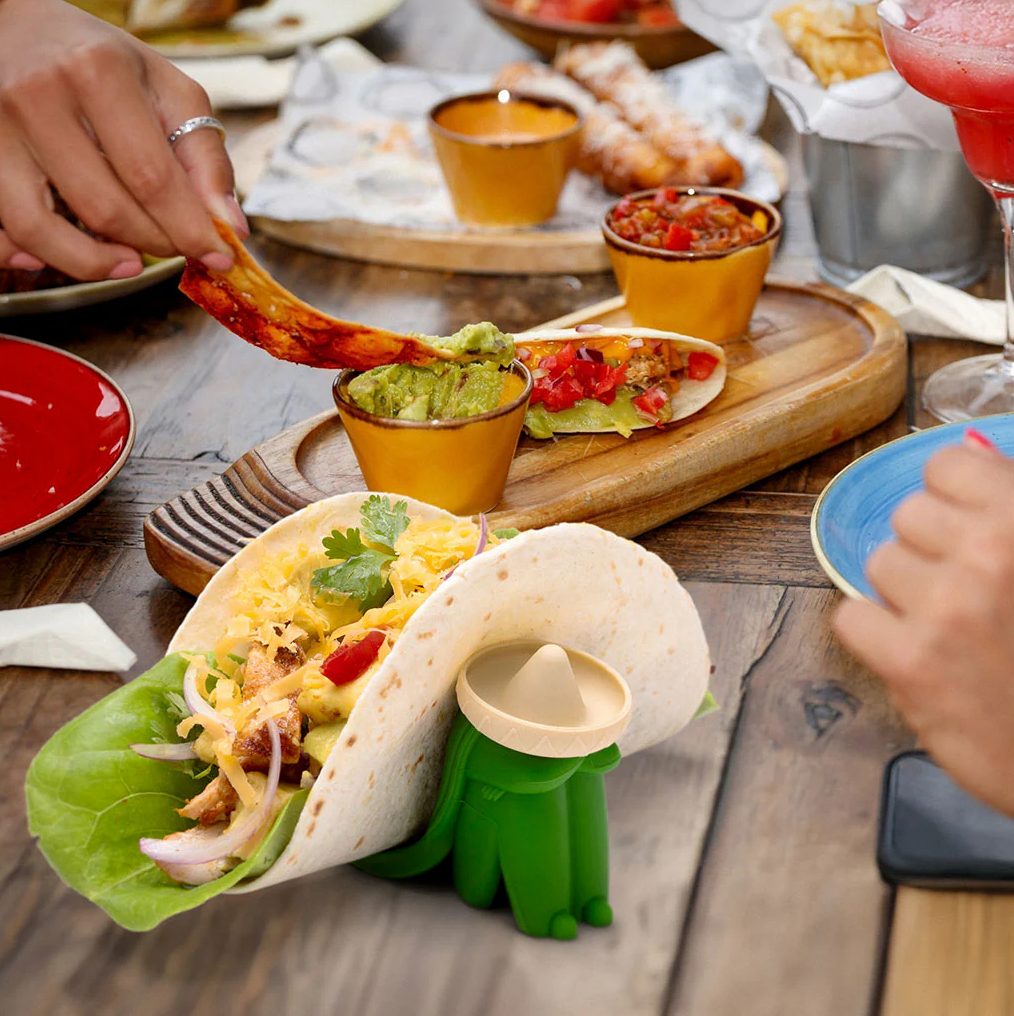 Monkey Business Los Dos Taco Holders. Person holding a taco shell with a small green alien figure inside, surrounded by food on a wooden table.