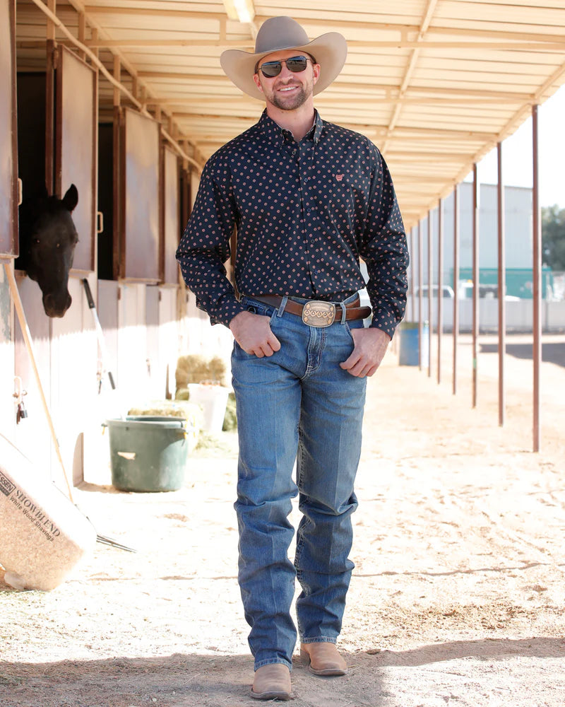 Man in cowboy attire standing in a stable with a horse in the background