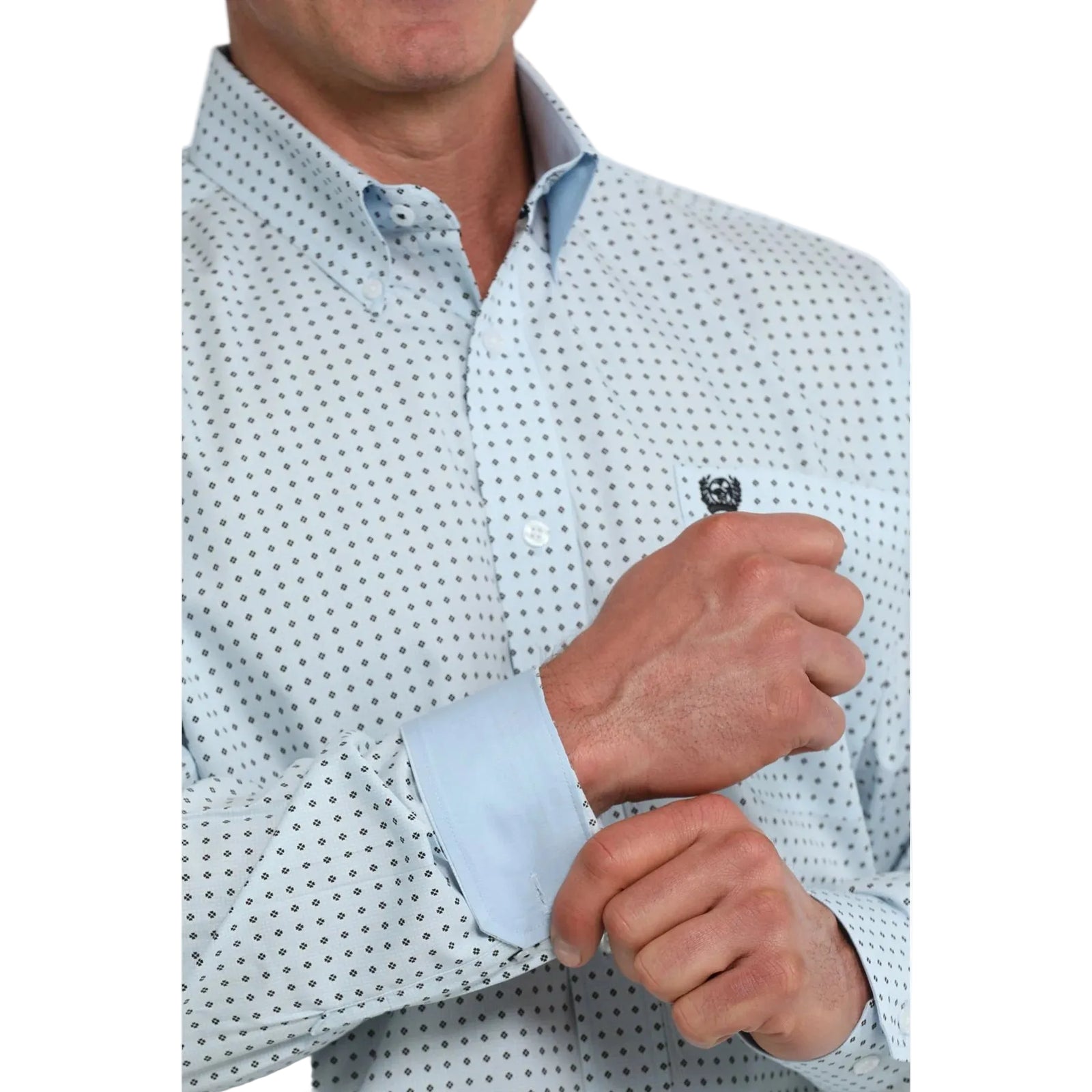 A close-up of a man wearing a light blue long-sleeve button-down shirt with a small black geometric dot pattern and a light blue solid inner collar, adjusting his cuffs.