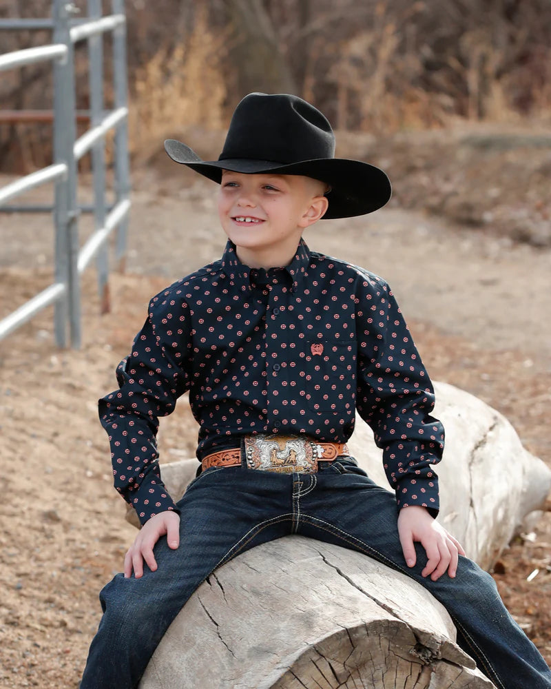 Child wearing a cowboy hat and patterned shirt sitting on a log outdoors.