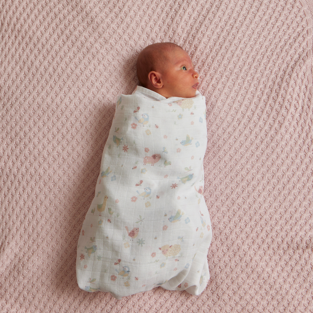 Jiggle & Giggle Birdy Muslin Wrap. Newborn baby swaddled in a patterned blanket on a textured pink surface