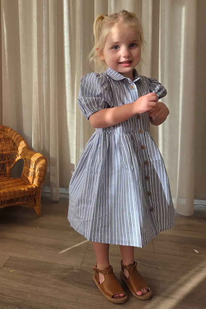 Milky Girl's Marine Stripe Dress. Young girl in a striped dress standing indoors with curtains and a chair in the background.