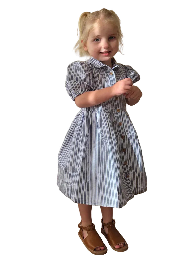 Milky Girl's Marine Stripe Dress. Young girl in a striped dress standing indoors with curtains and a chair in the background.