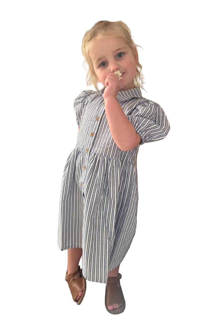 Milky Girl's Marine Stripe Dress. Young girl in a striped dress standing indoors with a chair and curtains in the background.