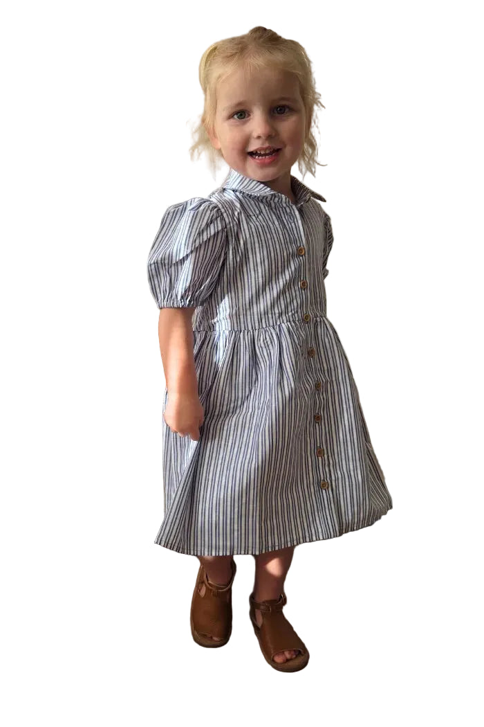 Milky Girl's Marine Stripe Dress. Young girl in a striped dress standing in a room with curtains and a wicker chair.
