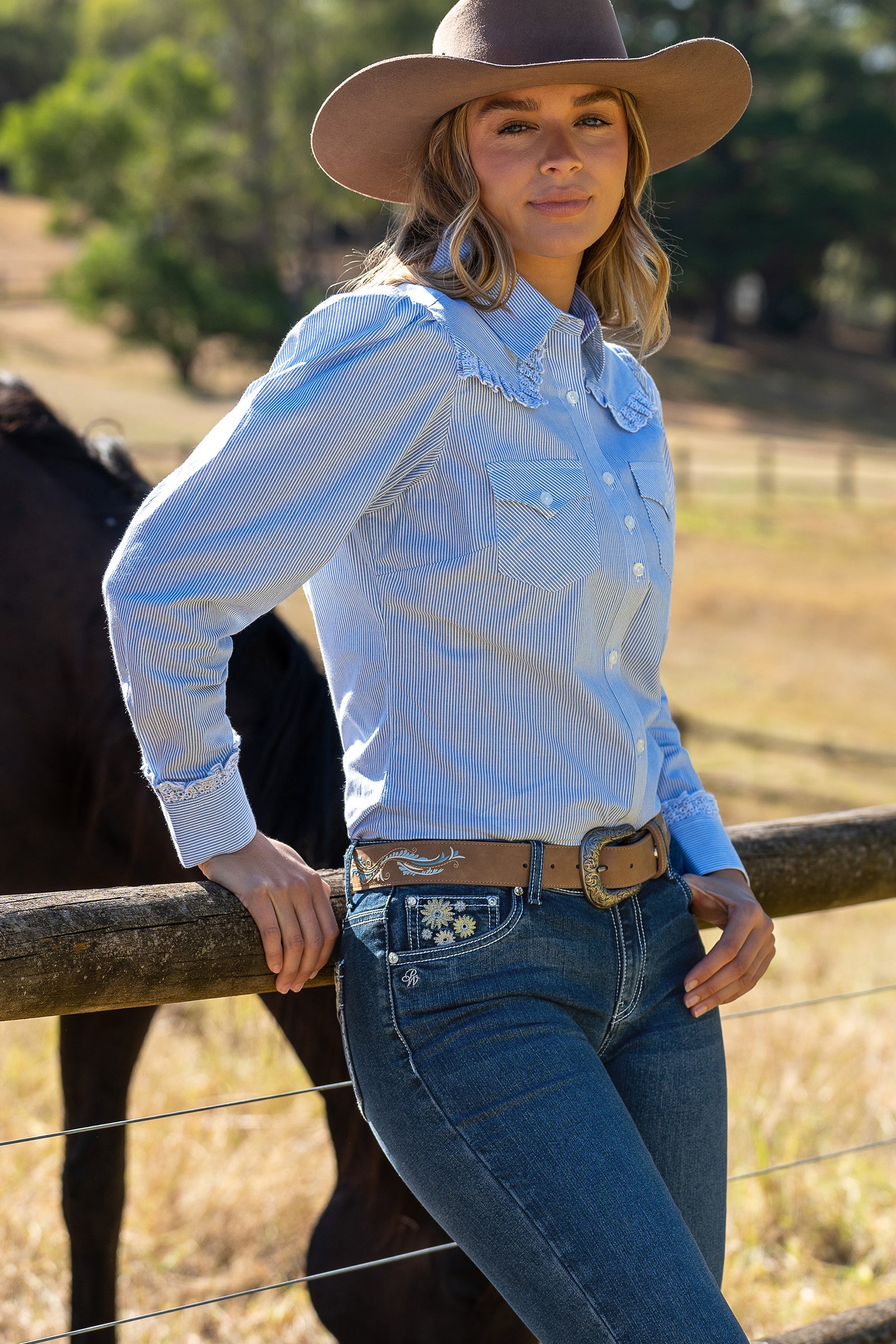 Pure Western Women's Lexi L/S Western Shirt. Woman in a light blue shirt and cowboy hat standing next to a horse in a field.