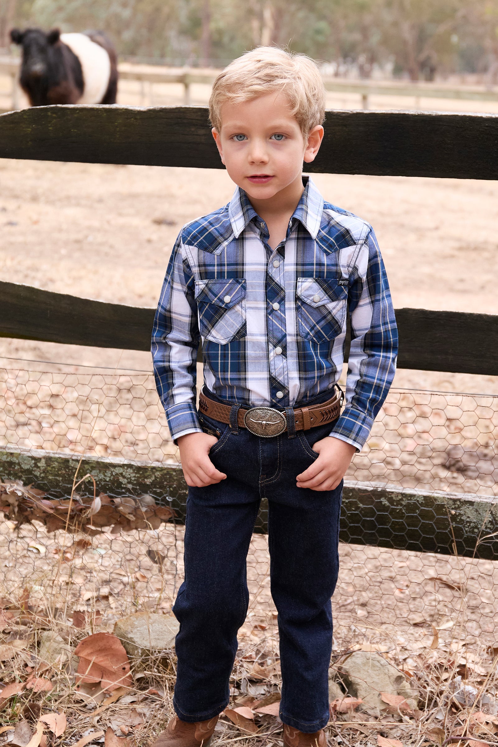 Pure Western Boy's Nicholas L/S Western Shirt. Young boy in a plaid shirt and jeans standing in front of a wooden fence.