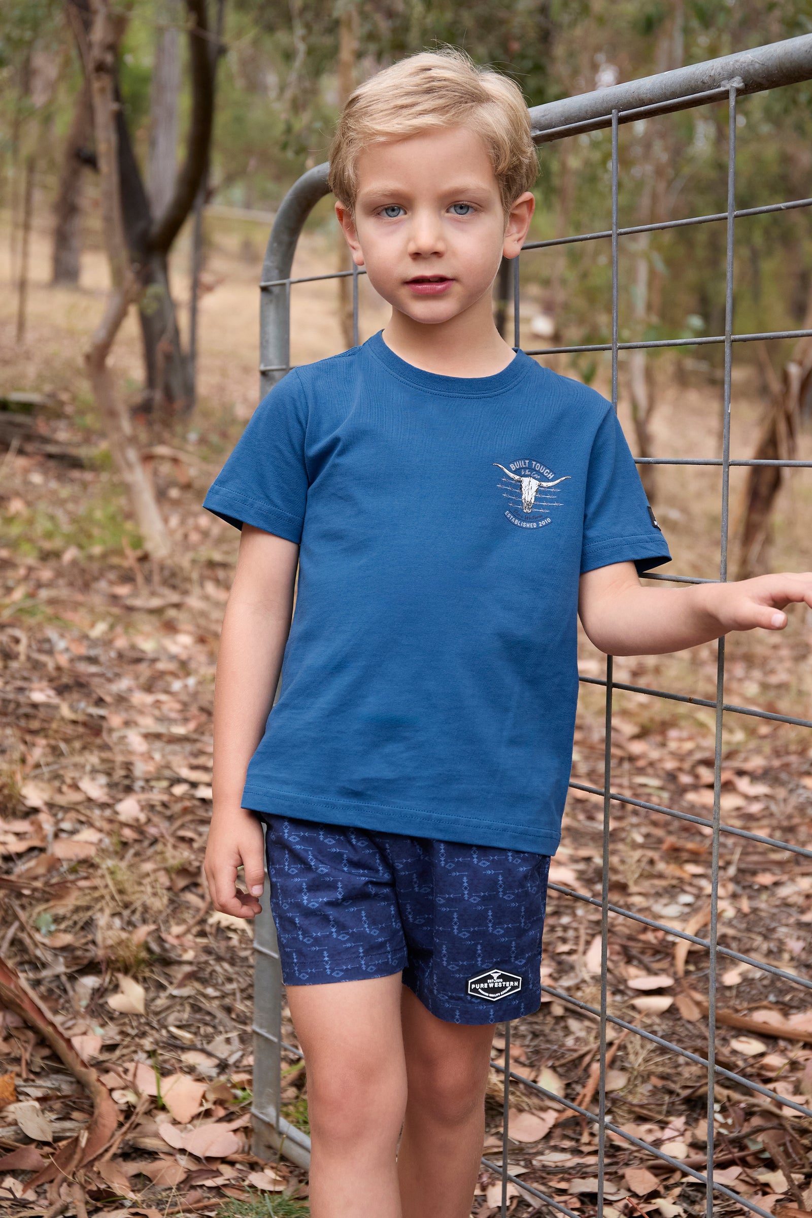Pure Western Boy's Frank S/S T-Shirt. Young boy wearing a blue t-shirt and shorts standing near a wire fence outdoors.