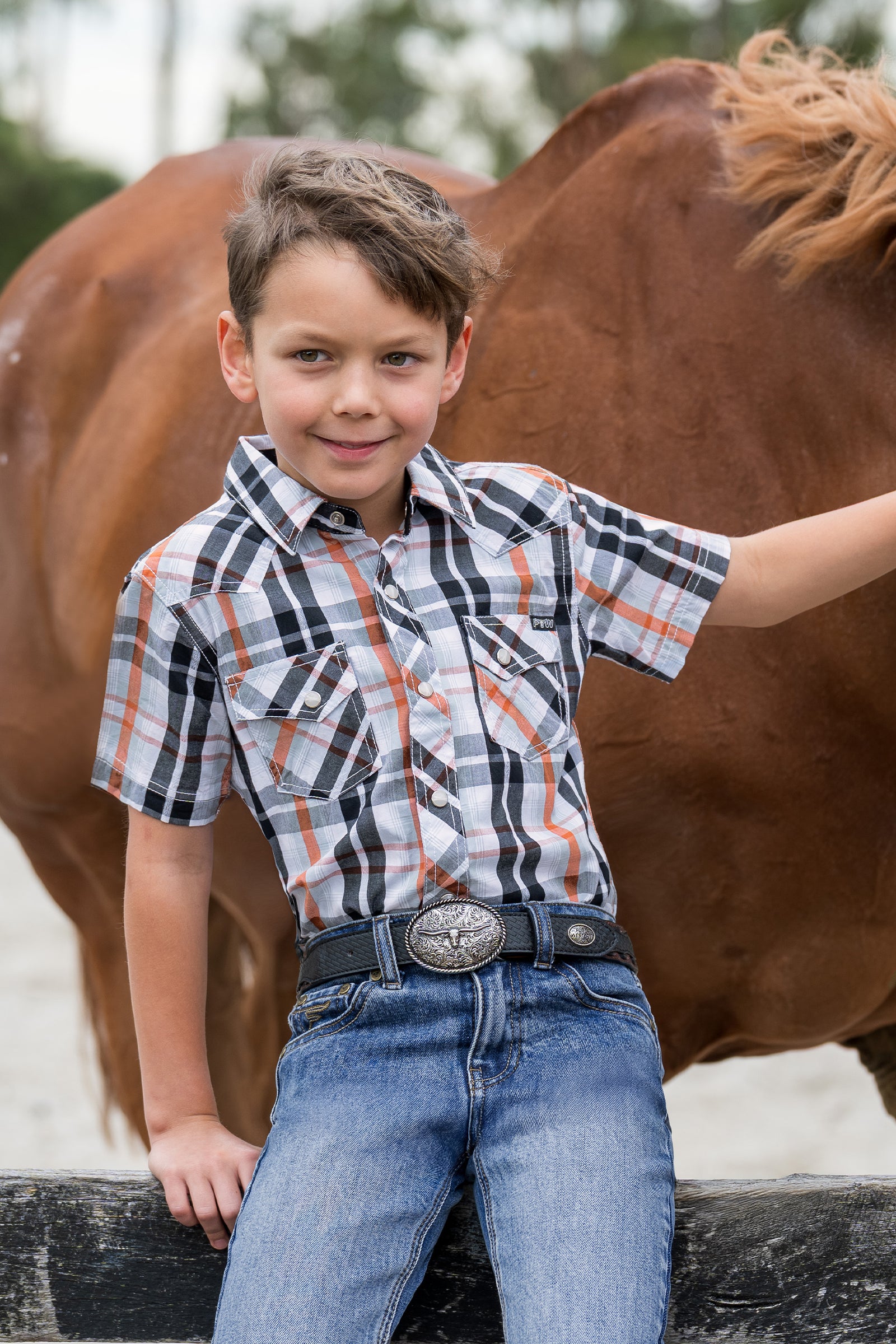 A young boy wearing a western-style plaid short-sleeve shirt, blue jeans, and a silver concho belt, posing in front of a brown horse.