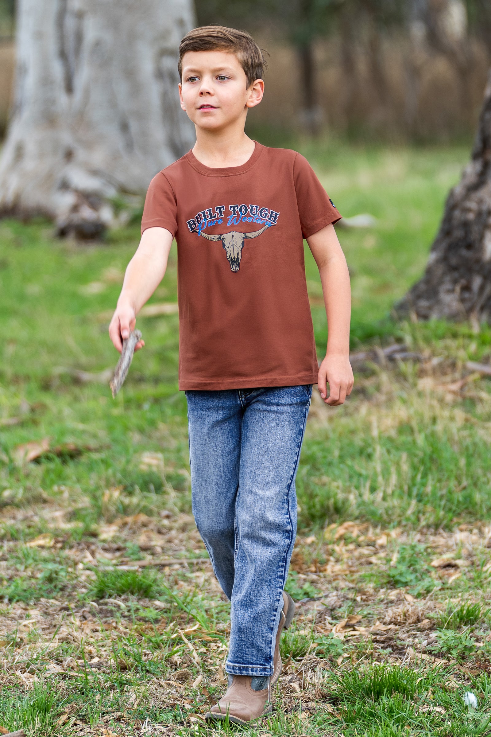 A young boy wearing a rust brown graphic t-shirt with 'RUSTIC TOUGH' and a steer skull design, paired with blue jeans, walking outdoors on a grassy path.