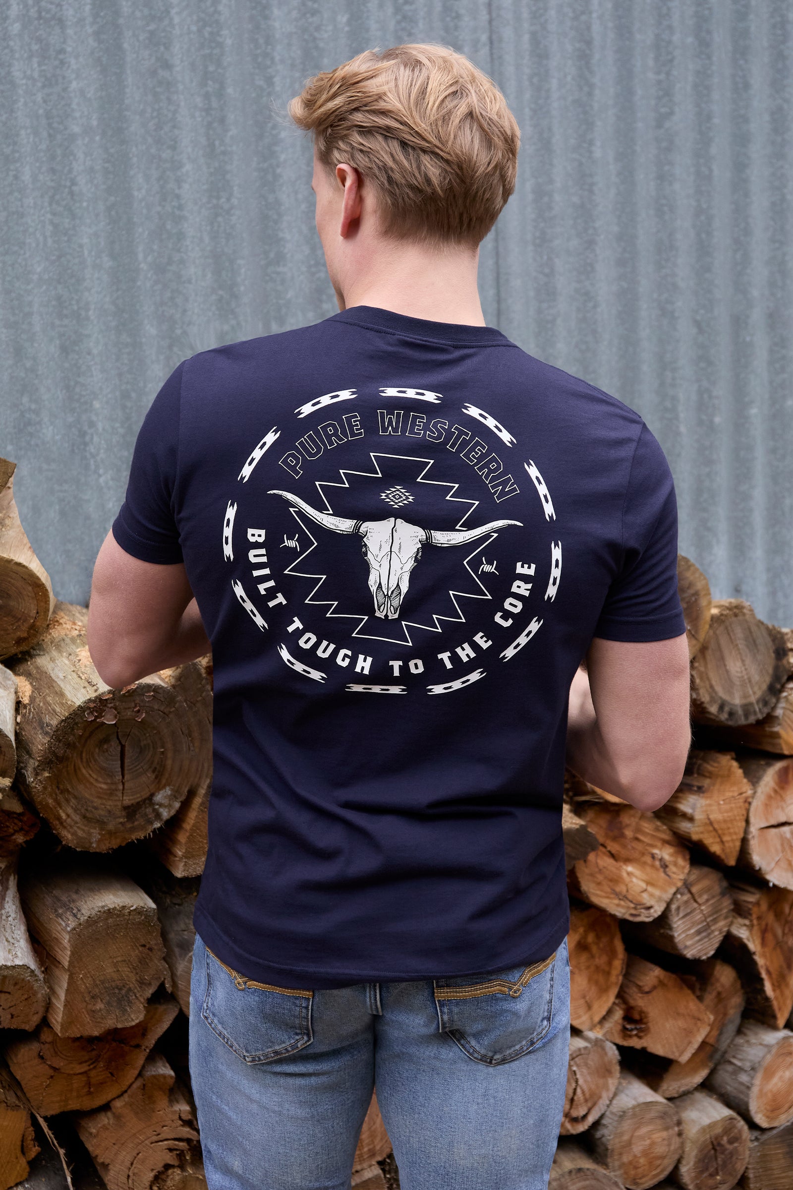 Man wearing a navy blue t-shirt with a graphic design, standing in front of stacked logs.